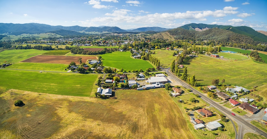 An aerial view of a rural town with green fields, houses, and mountains in the background.