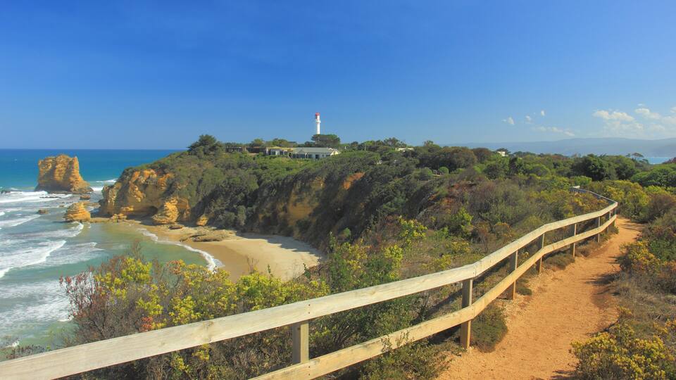 A coastal landscape with a lighthouse, rocky cliffs, and a wooden walkway along the beach.