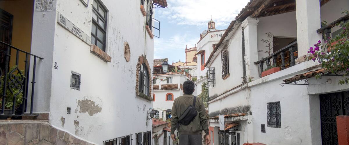 A narrow street with white buildings, a man walking, and a hotel in the background.