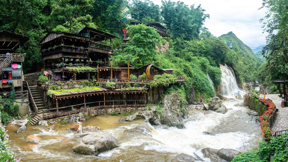 A river with a waterfall, wooden buildings, and lush greenery.