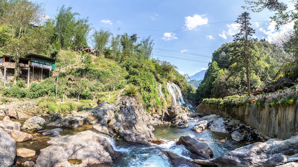 A river flowing through a mountainous area with a waterfall, surrounded by dense greenery and traditional houses.