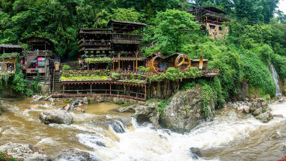 A river with a waterfall, surrounded by lush greenery and traditional wooden structures.