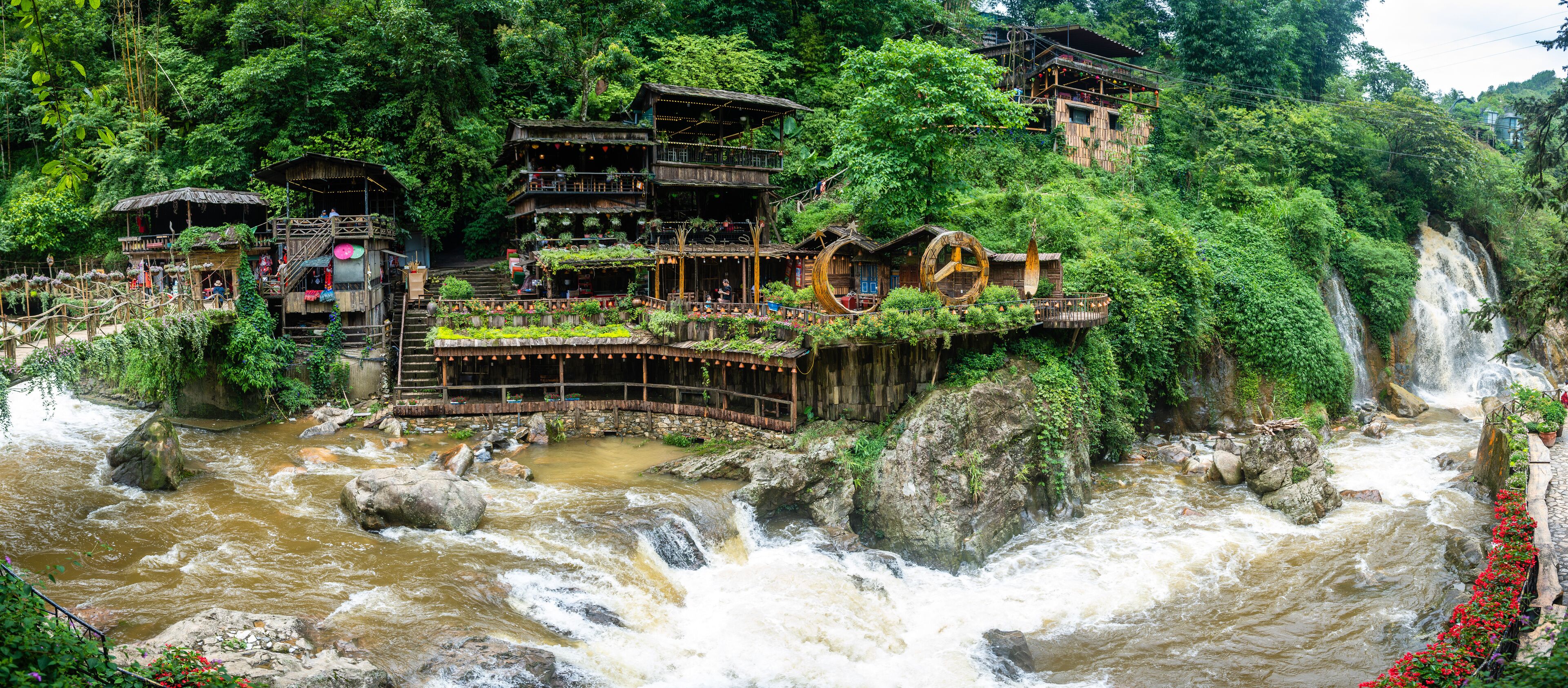 A river with a waterfall, surrounded by lush greenery and traditional wooden structures.