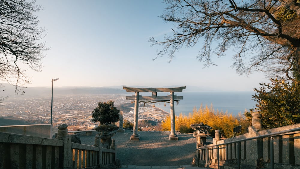 A traditional Japanese torii gate at the top of a hill overlooking a cityscape.