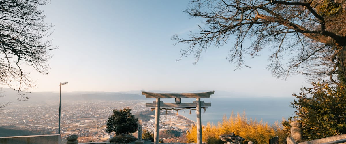View of a Torii gate standing majestically against the backdrop of the sea and clear blue sky, framed by bare tree branches, Kanonji, Kagawa, Japan.