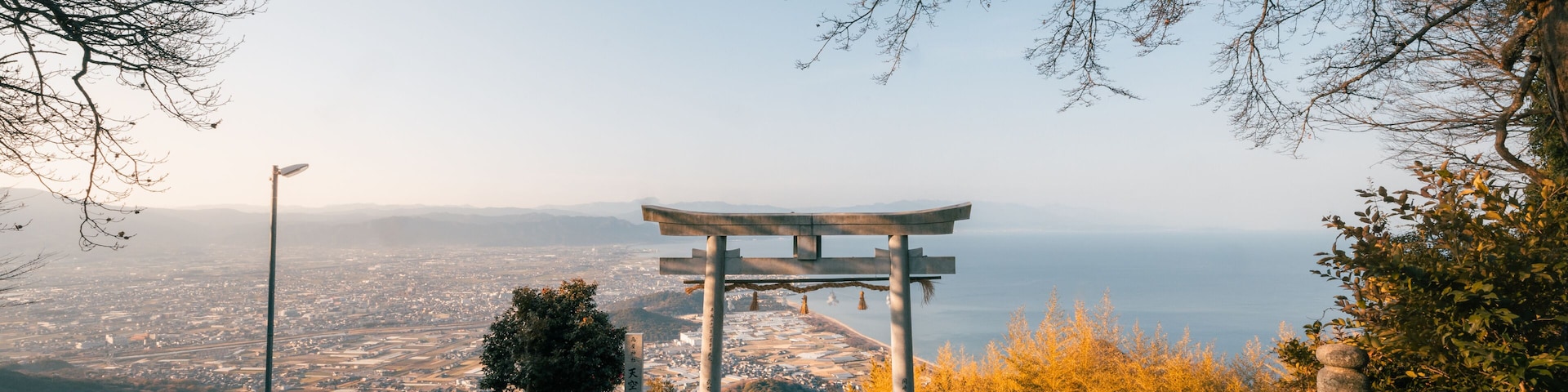 View of a Torii gate standing majestically against the backdrop of the sea and clear blue sky, framed by bare tree branches, Kanonji, Kagawa, Japan.