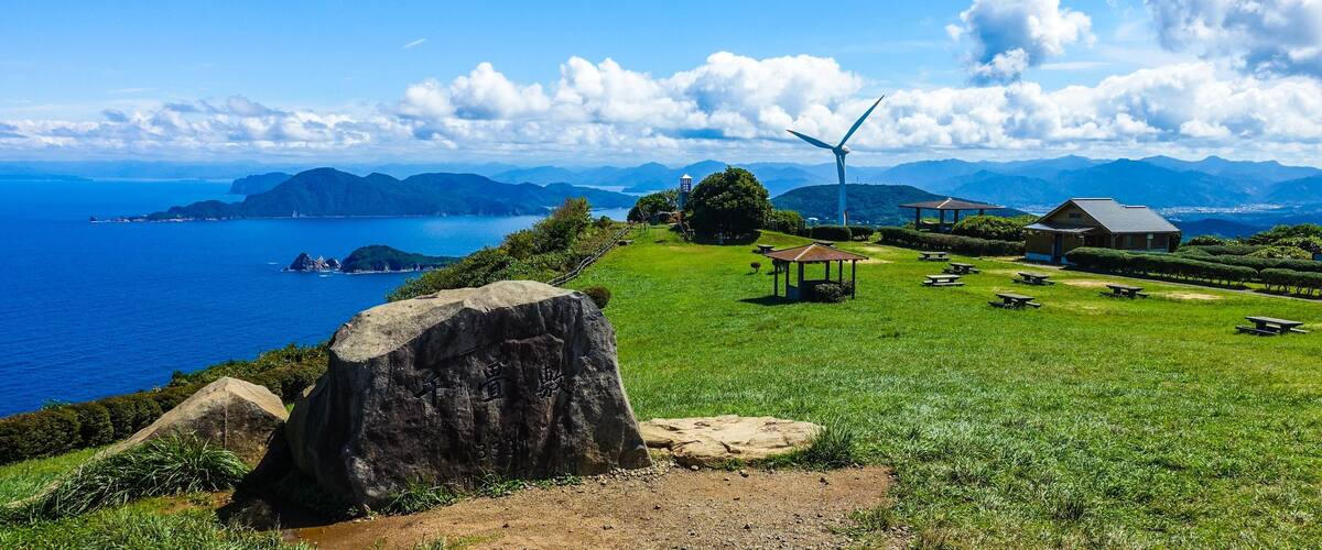 A coastal landscape with a wind turbine, picnic areas, and a large rock with inscriptions.