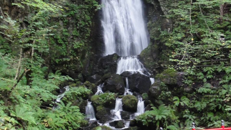 A waterfall cascading down a rocky cliff surrounded by lush greenery. A red bridge with a railing is visible in the foreground.