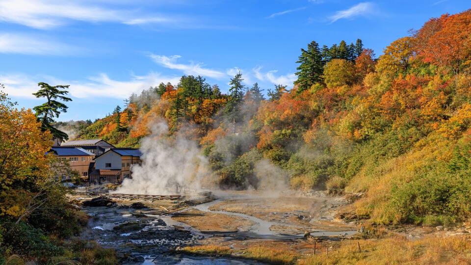 A geothermal area with steam vents, a river, and autumn-colored trees.