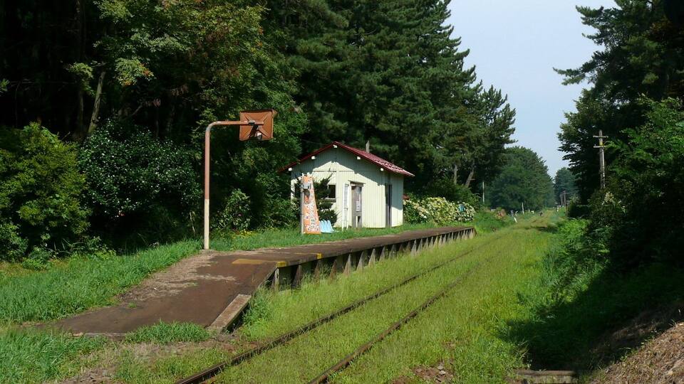 A rural railway station with a small building, tracks, and surrounding greenery.