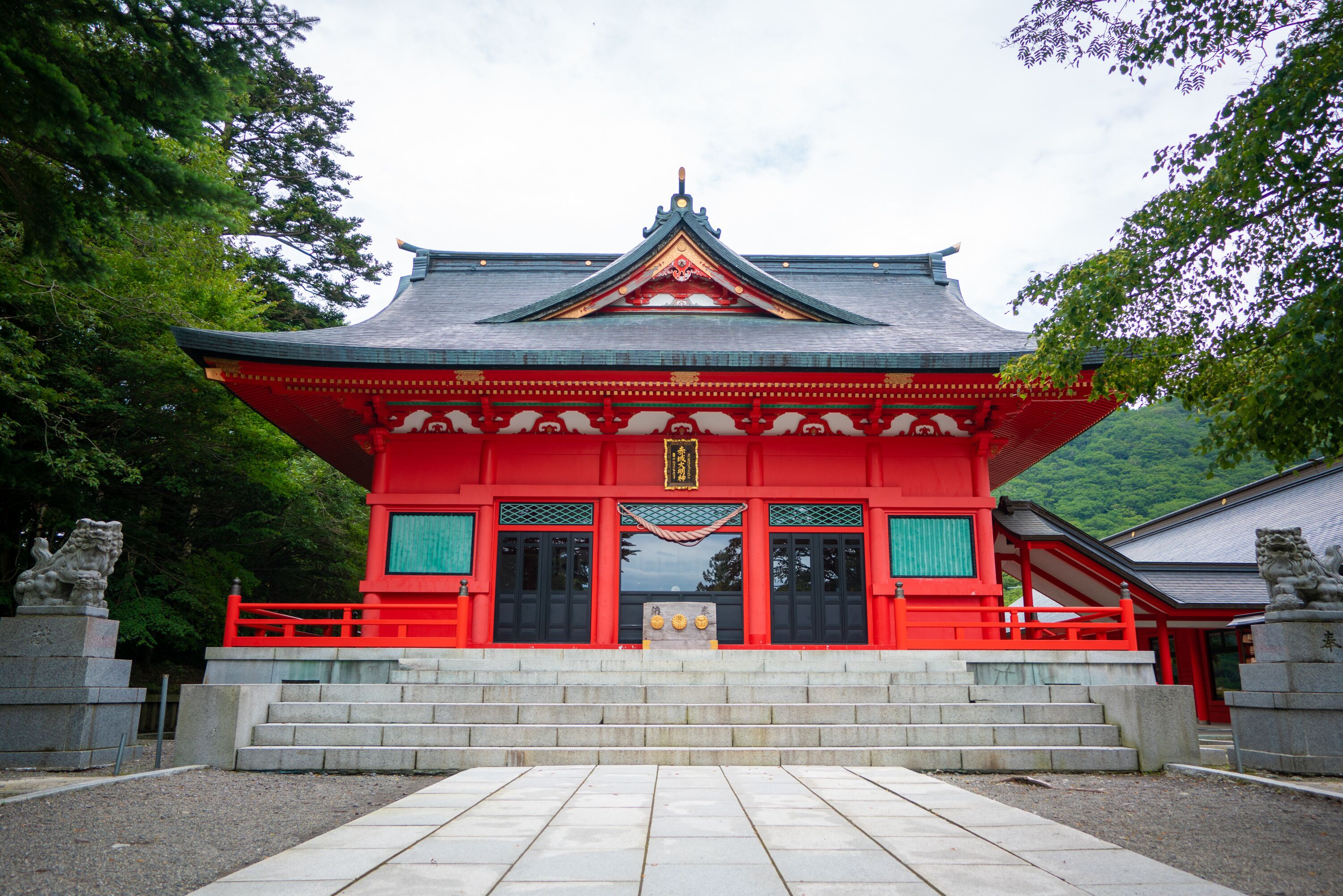 A traditional Japanese temple with a red roof and grey walls, surrounded by trees.