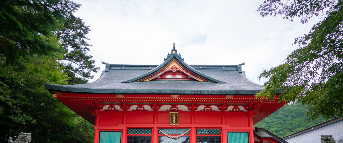 A traditional Japanese temple with a red roof and grey walls, surrounded by trees.