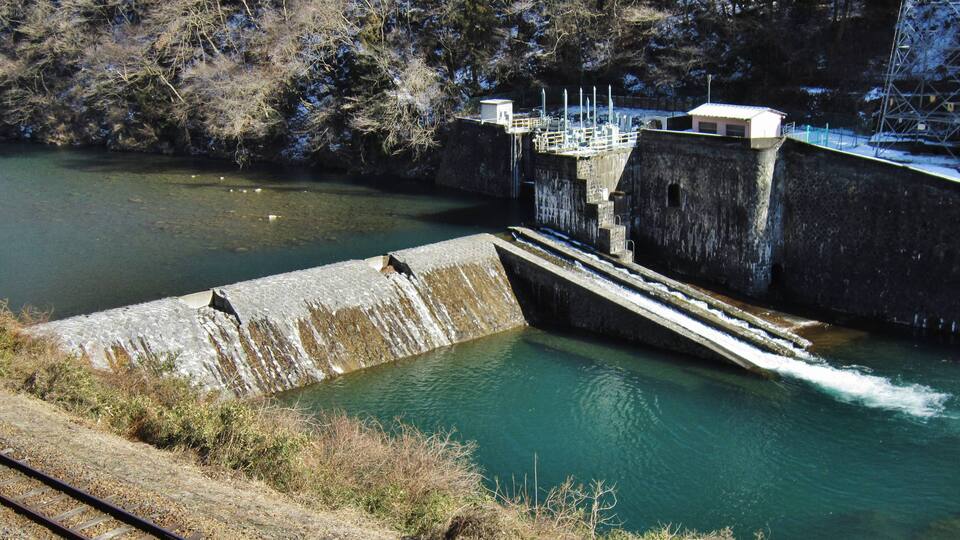 A dam with water flowing over it, surrounded by trees and a railway track.