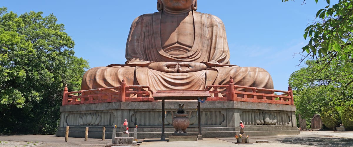A large seated Buddha statue with a railing, surrounded by trees and a clear sky.