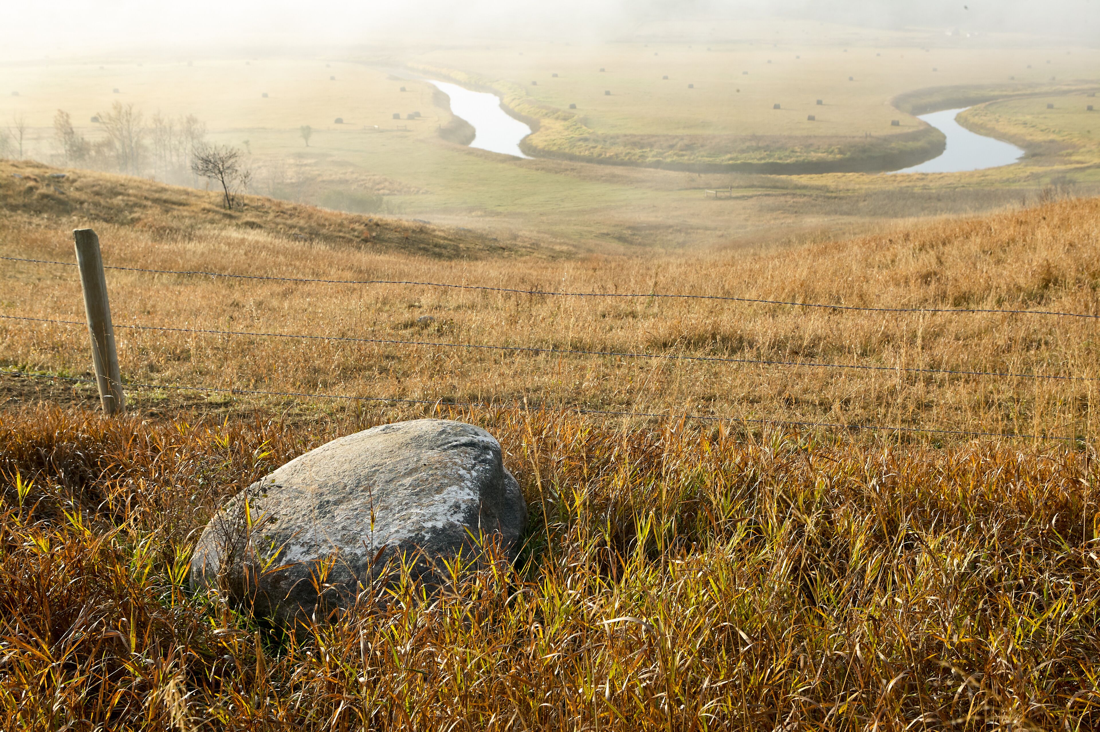 A grassy field with a large rock in the foreground and a winding river in the background.