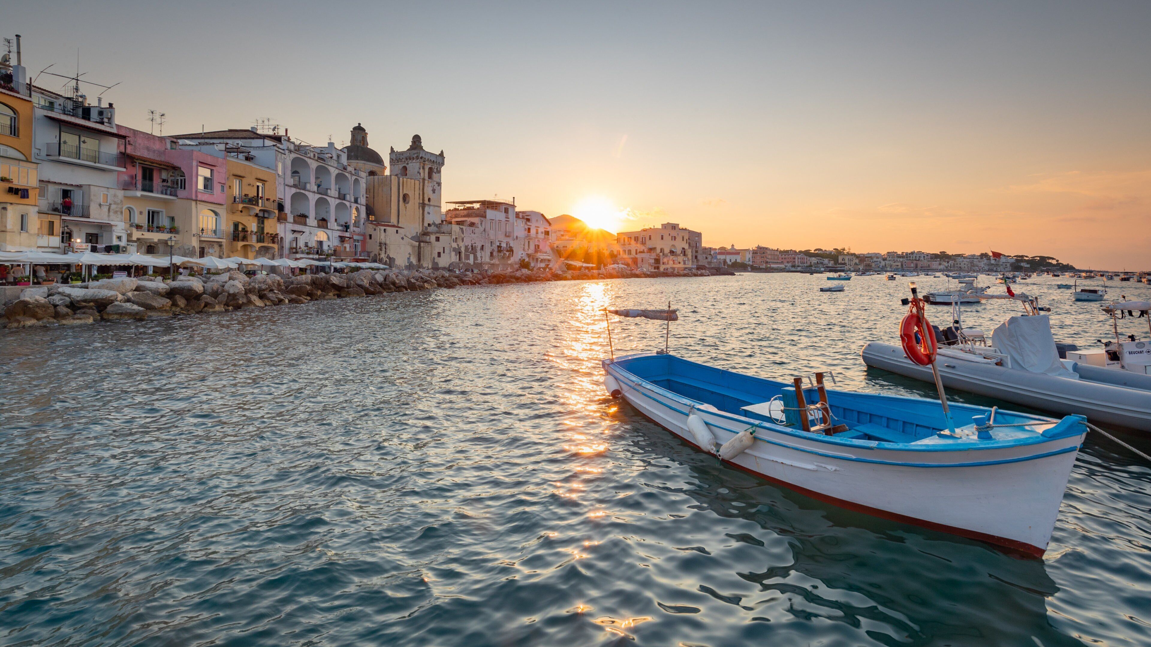 A coastal town with colorful buildings, a calm sea, and boats docked at the harbor during sunset.