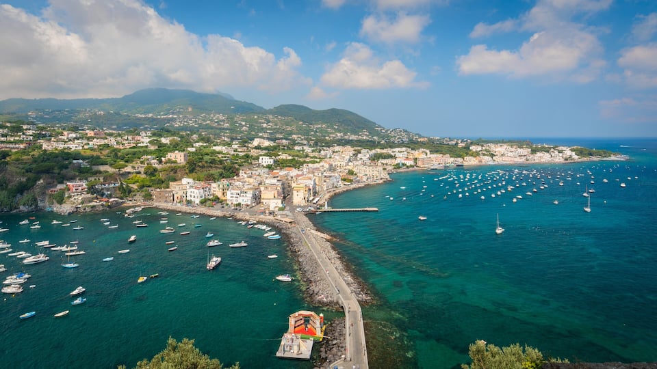 A coastal town with a harbor filled with boats, a rocky breakwater, and a backdrop of mountains.