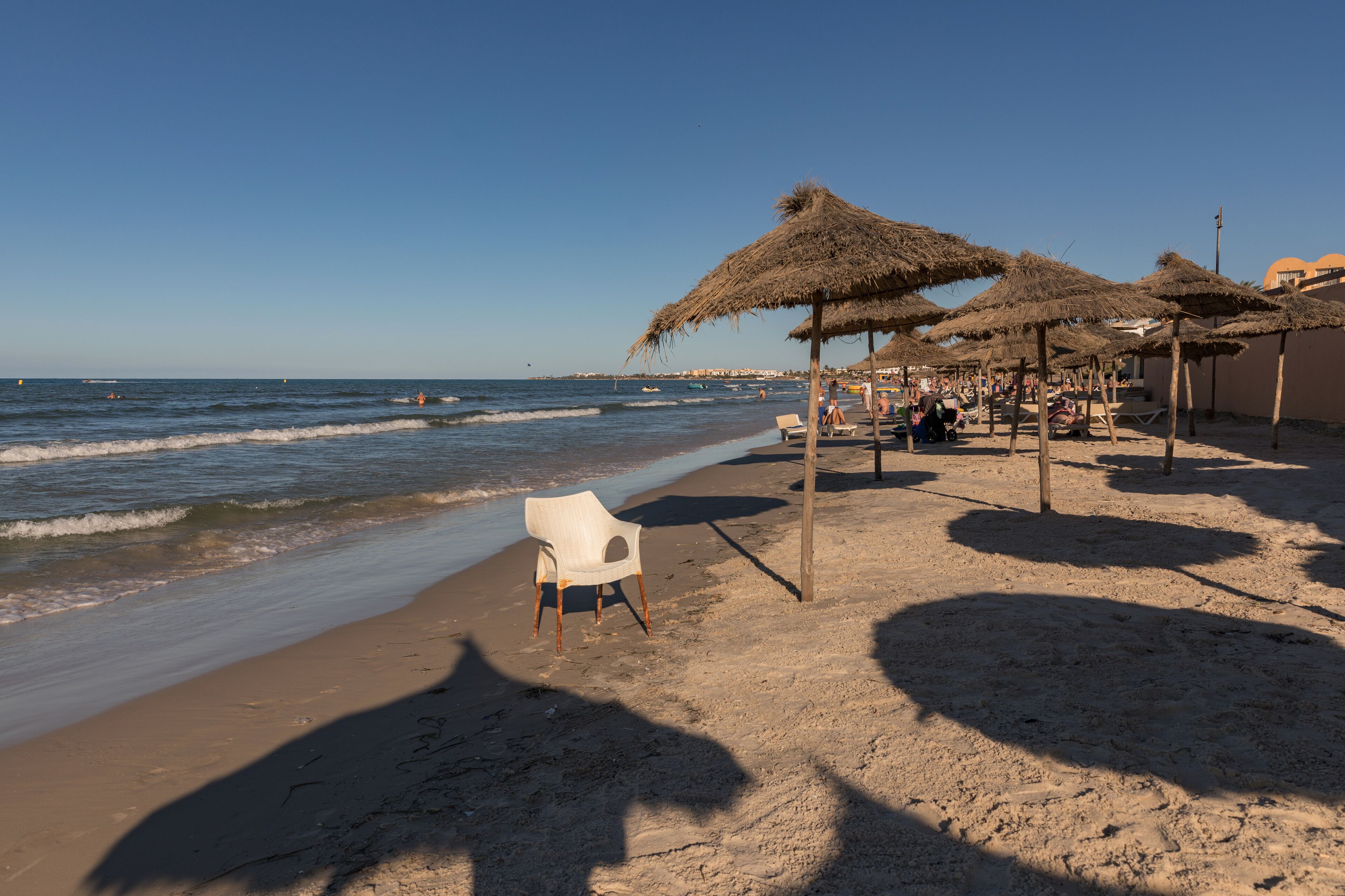 A beach with thatched umbrellas, a white plastic chair, and a clear blue sky.