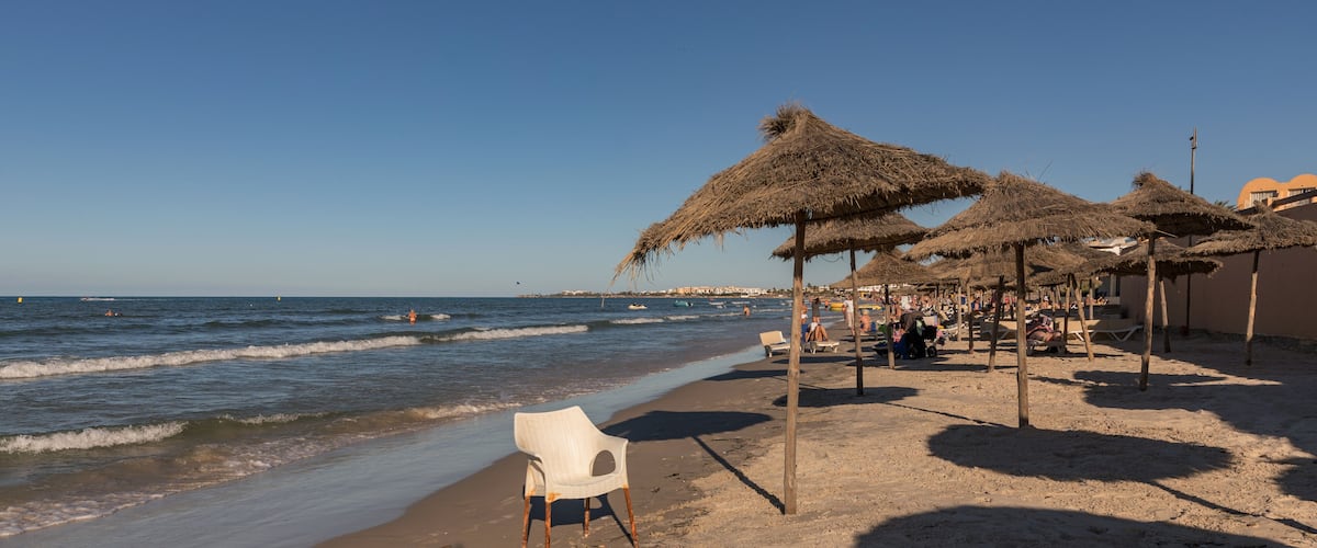A beach with thatched umbrellas, a white plastic chair, and a clear blue sky.