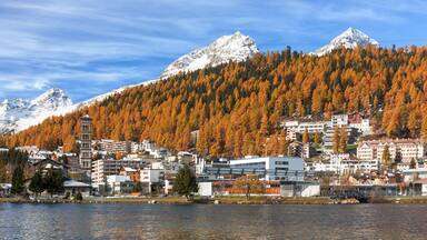 A town by a lake with autumn-colored trees, snow-capped mountains, and a mix of modern and traditional buildings.