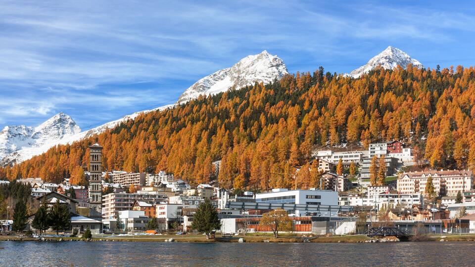 A town by a lake with autumn-colored trees, snow-capped mountains, and a mix of modern and traditional buildings.