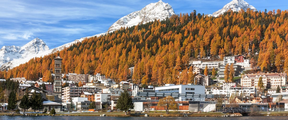 A town by a lake with autumn-colored trees, snow-capped mountains, and a mix of modern and traditional buildings.