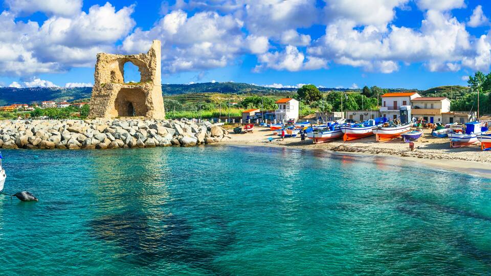 A coastal town with a historic stone structure, boats, and a clear blue sea.