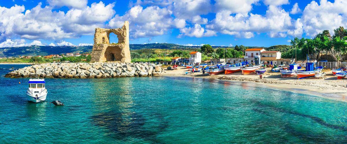 A coastal town with a historic stone structure, boats, and a clear blue sea.