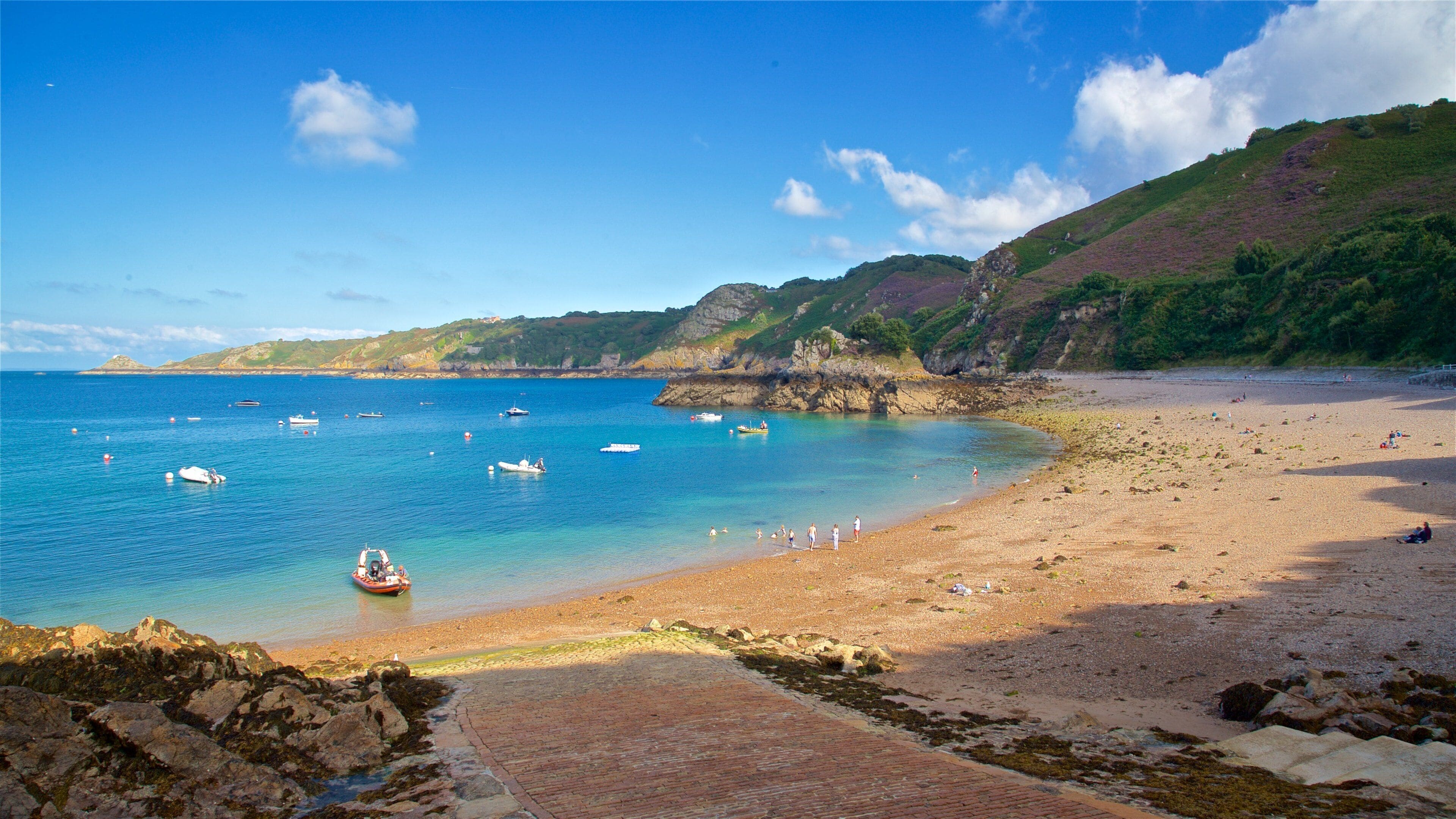 A coastal beach with clear blue waters, boats, and rocky cliffs.