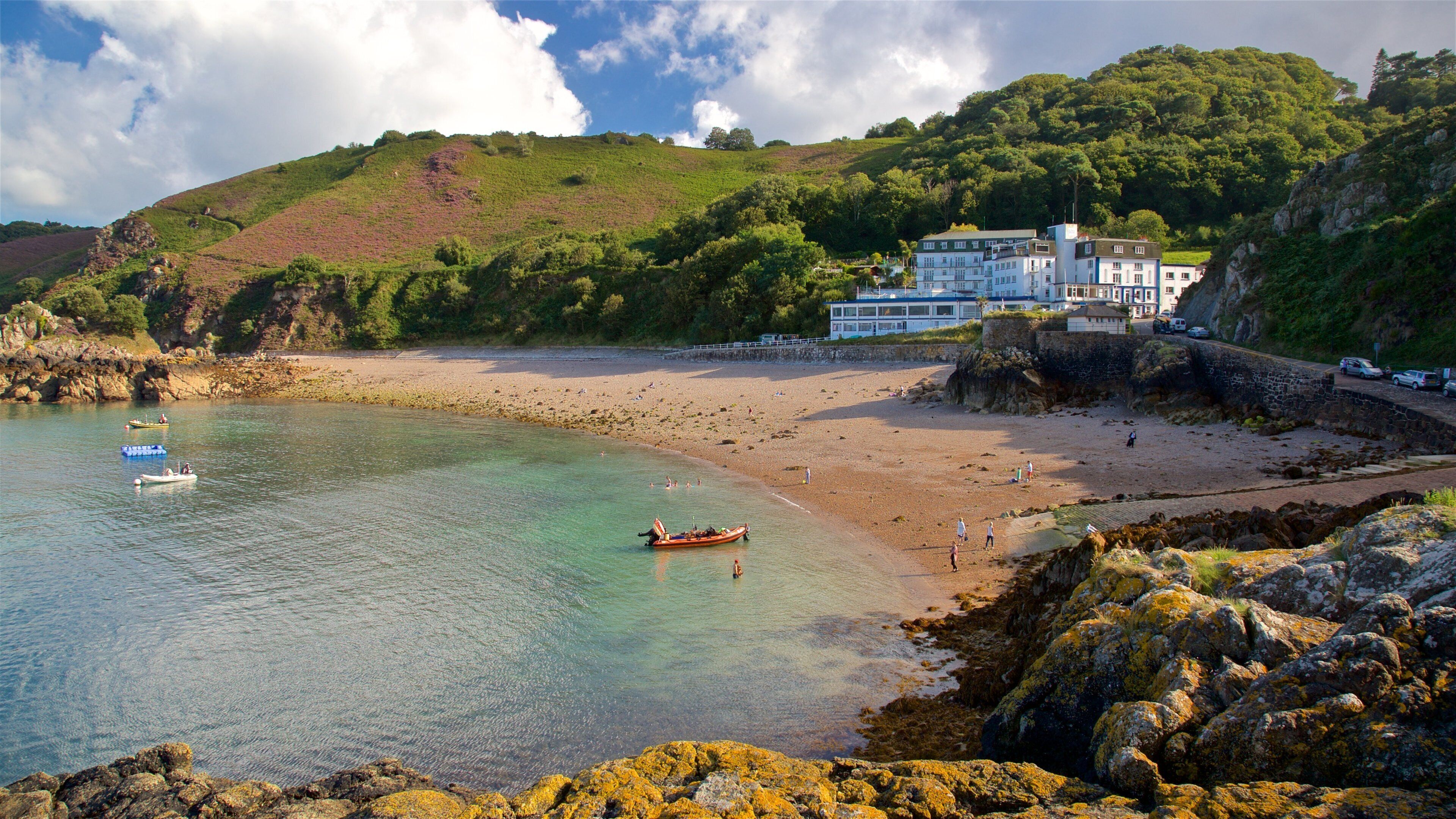 A coastal landscape with a sandy beach, clear water, rocky shore, and a hill with vegetation.