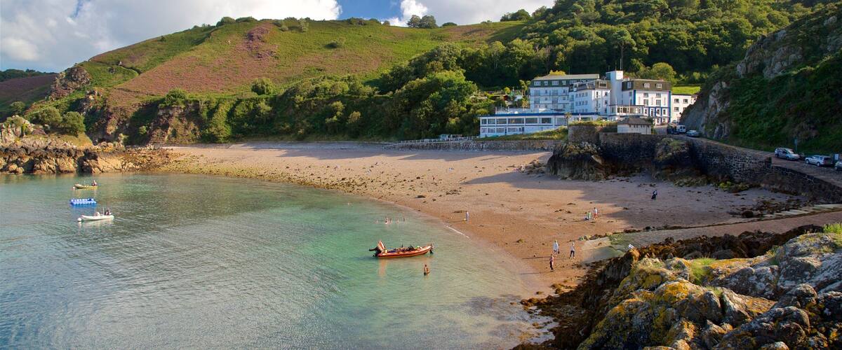 A coastal landscape with a sandy beach, clear water, rocky shore, and a hill with vegetation.