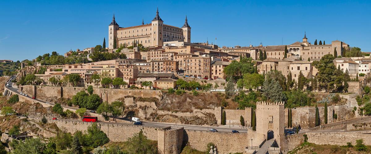 A historic city with a prominent castle on a hill, surrounded by ancient walls and a winding road.