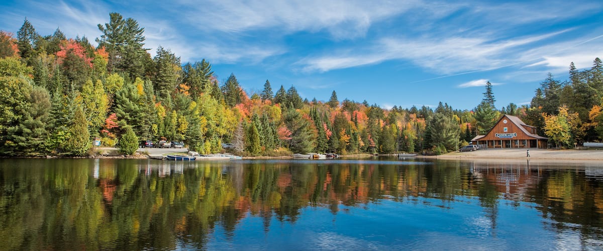 A calm lake with a wooden dock, surrounded by autumn-colored trees and a lodge in the background.