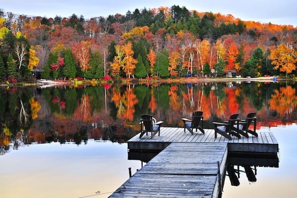 A calm lake with autumn-colored trees reflecting on the water, a wooden dock with chairs, and a small cabin in the background.
