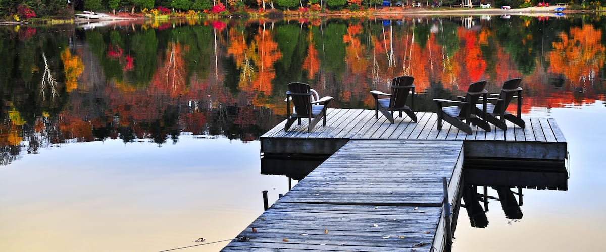 A calm lake with autumn-colored trees reflecting on the water, a wooden dock with chairs, and a small cabin in the background.