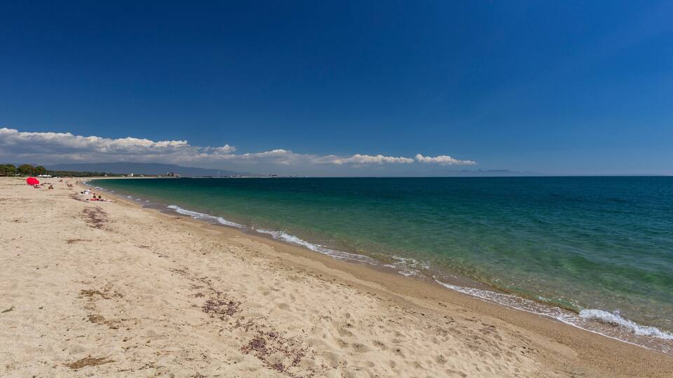 A sandy beach with clear turquoise water, a few people, and distant mountains under a blue sky.