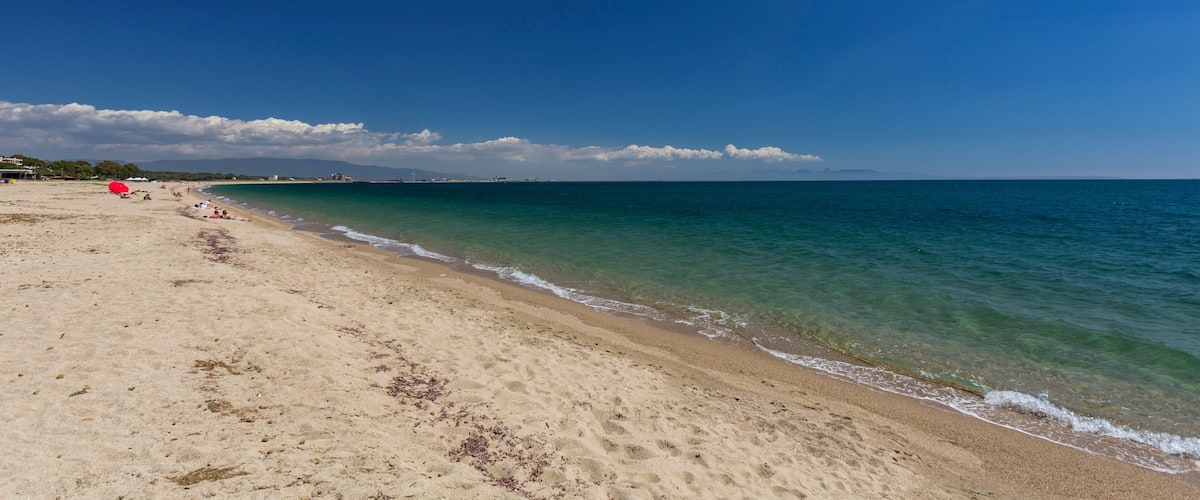 A sandy beach with clear turquoise water, a few people, and distant mountains under a blue sky.