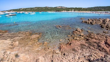 A coastal area with clear turquoise waters, rocky shorelines, boats, and a distant beach populated with people.