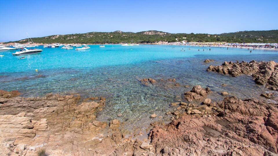A coastal area with clear turquoise waters, rocky shorelines, boats, and a distant beach populated with people.