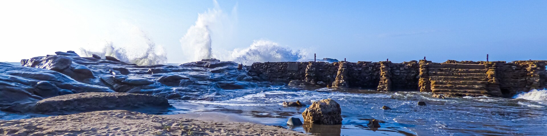 Uma costa rochosa com ondas quebrando contra um muro de pedra.