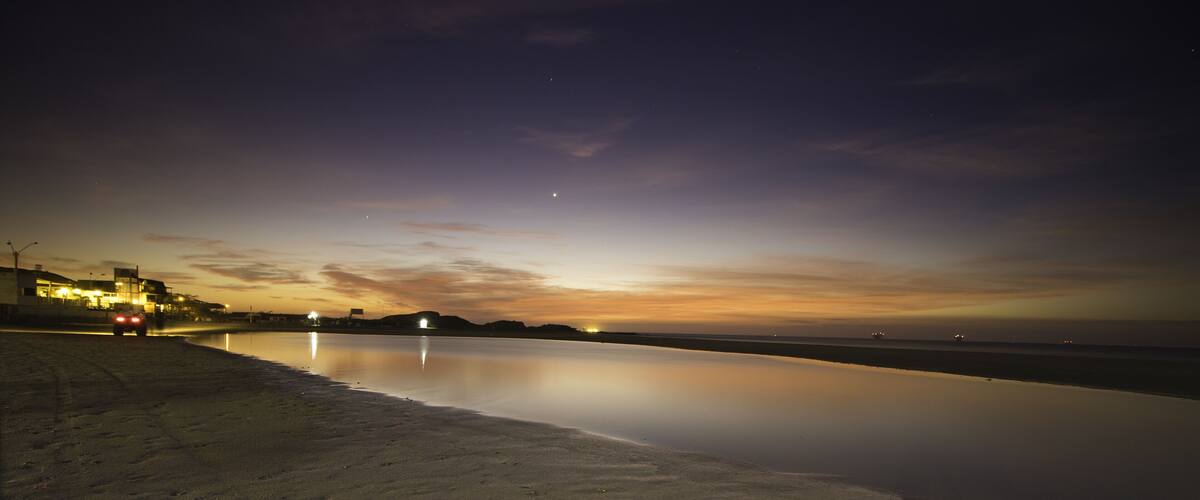 A beach at dusk with a calm sea, a distant city skyline, and a clear sky.