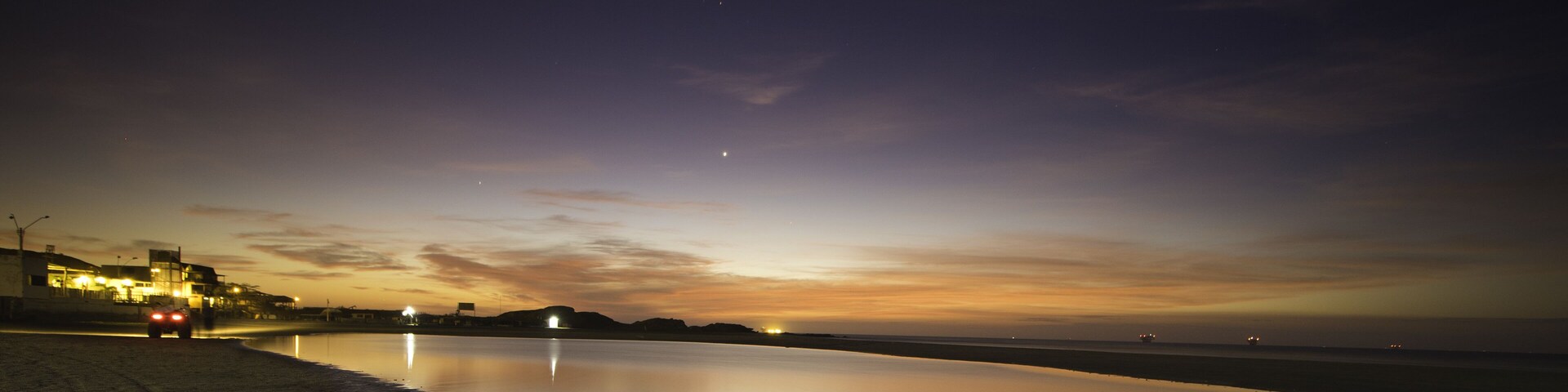 A beach at dusk with a calm sea, a distant city skyline, and a clear sky.