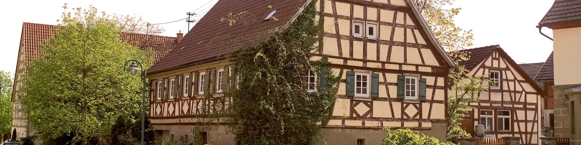 A traditional half-timbered house with a sloped roof, surrounded by greenery and a gravel driveway.