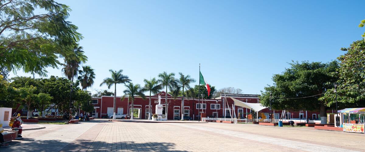 A plaza with a red building, palm trees, and a flag.