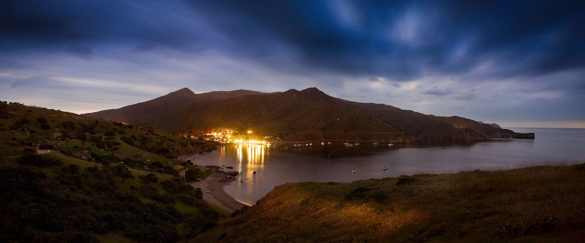 A coastal landscape with a mountain range, a lit-up village, and a calm sea.
