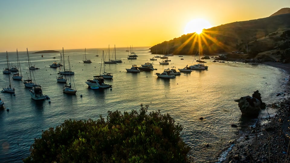 A coastal scene with boats anchored in calm waters, a rocky shoreline, and a sun setting in the background.