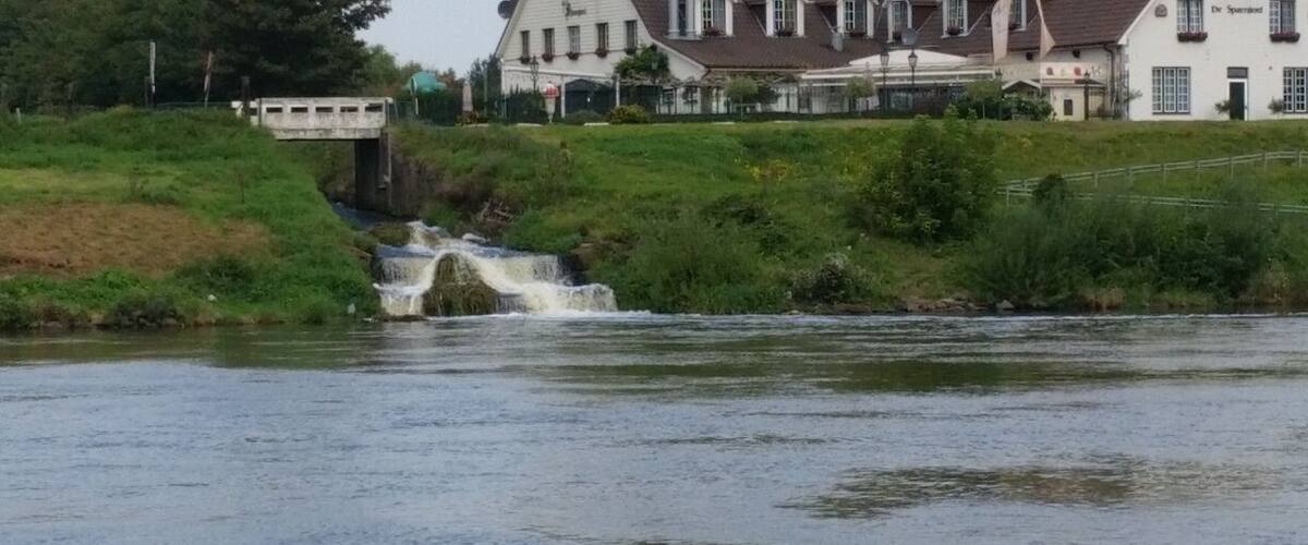 A river with a small waterfall and houses in the background.