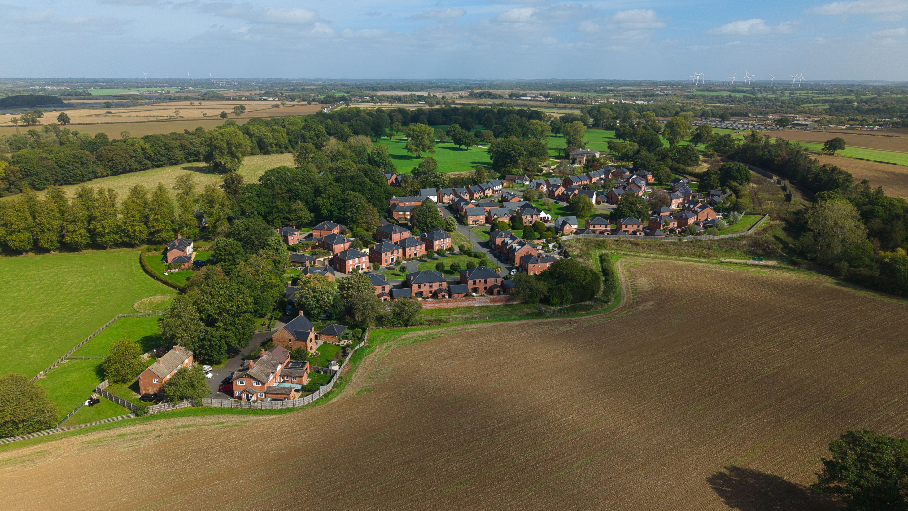 Un village rural avec des maisons, des champs et des éoliennes visibles au loin.