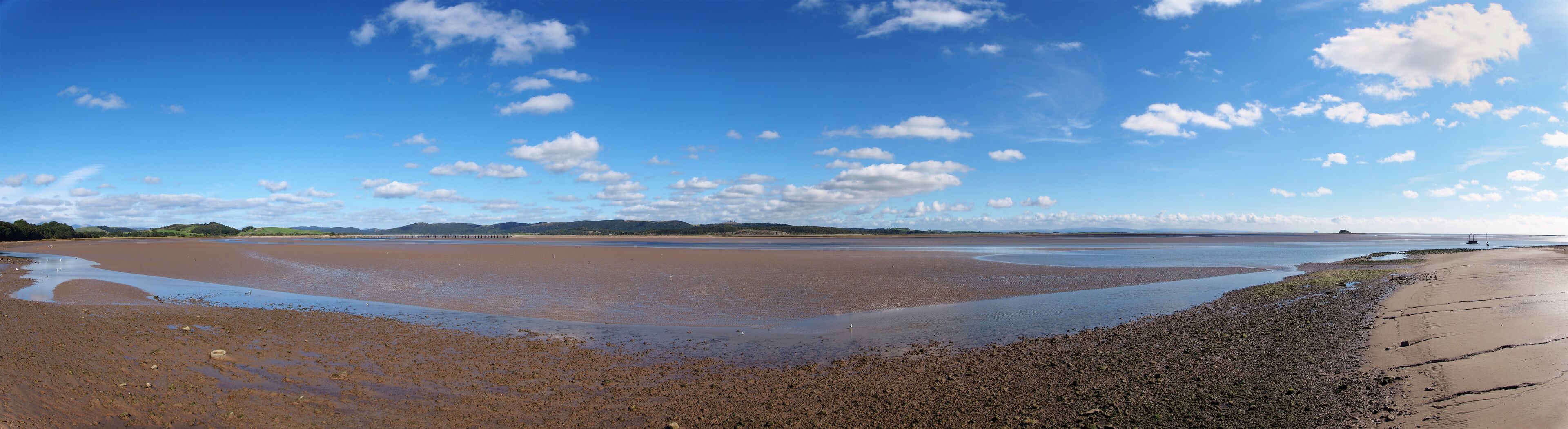 A coastal landscape with a wide, muddy beach, a clear blue sky, and distant hills.