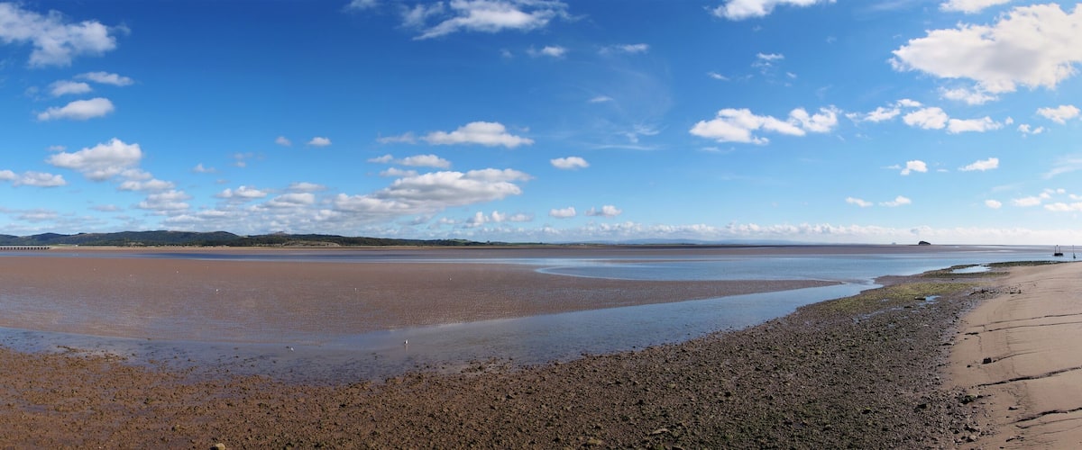 A coastal landscape with a wide, muddy beach, a clear blue sky, and distant hills.